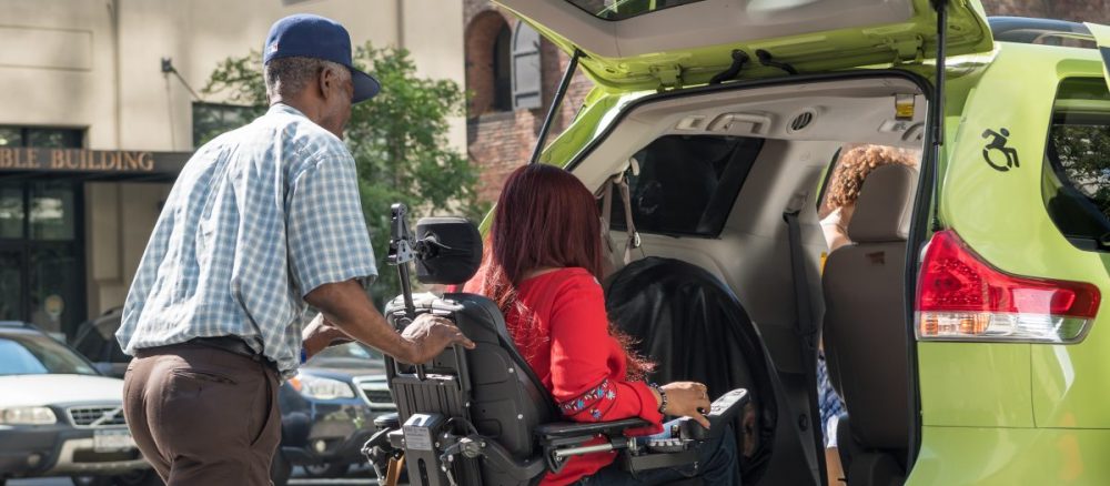 A taxi driver assists a woman who uses a motorized wheelchair into the back of a wheelchair accessible green taxi.