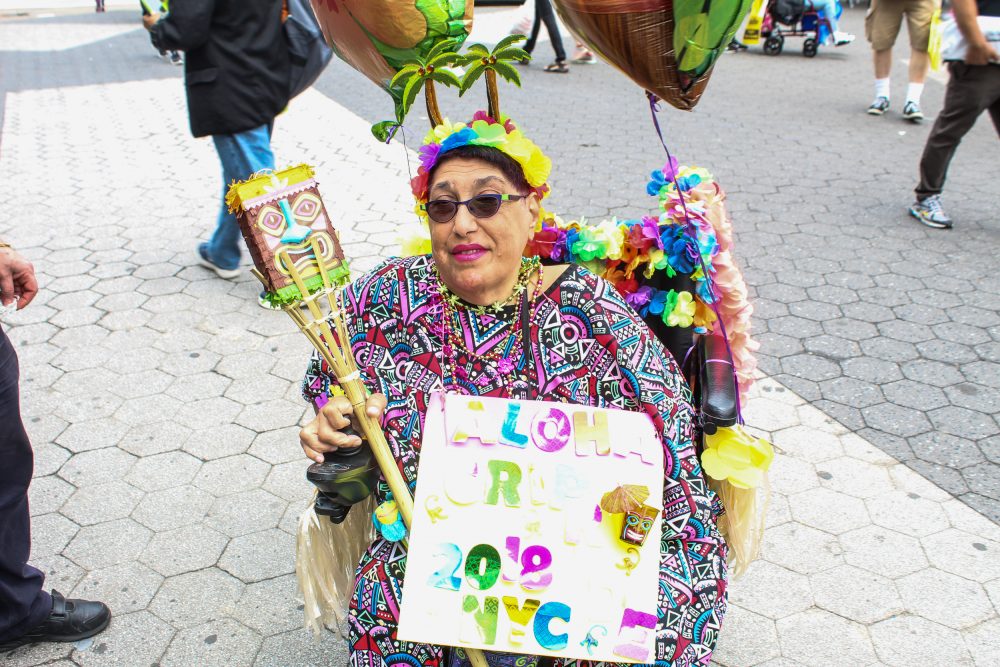 Woman in a wheelchair holds a sign and balloons while she celebrates at the Disability Pride Parade 2018 in colorful, festive attire.
