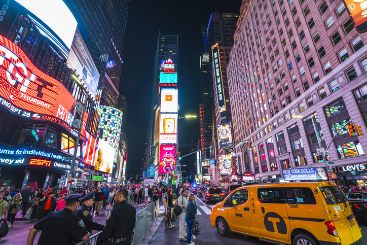 A photo shows a crowded Times Square at night time. A yellow wheelchair accessible taxi is shown in the foreground.