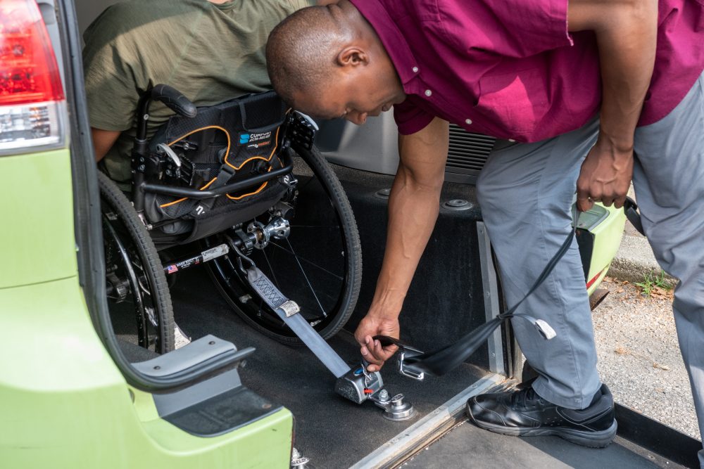 A driver secures his passenger's wheelchair in the rear of his green wheelchair accessible taxi using the securement straps.