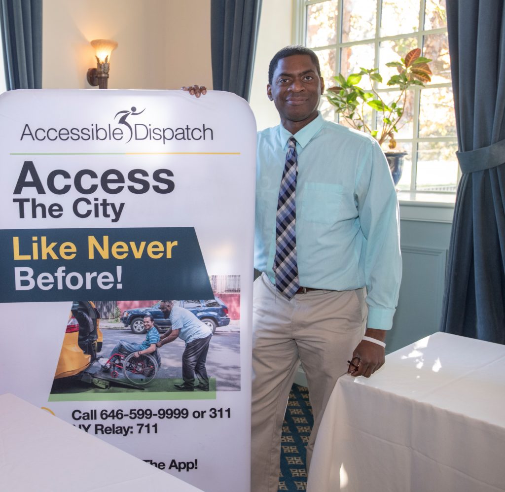 Accessible Dispatch Community Outreach Trainer Steven Williams stands beside a sign promoting the Accessible Dispatch program. The sign reads "Access the City Like Never Before."